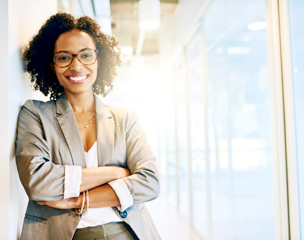 A young black woman in a stylish blazer and glasses smiles in a bright office hallway.